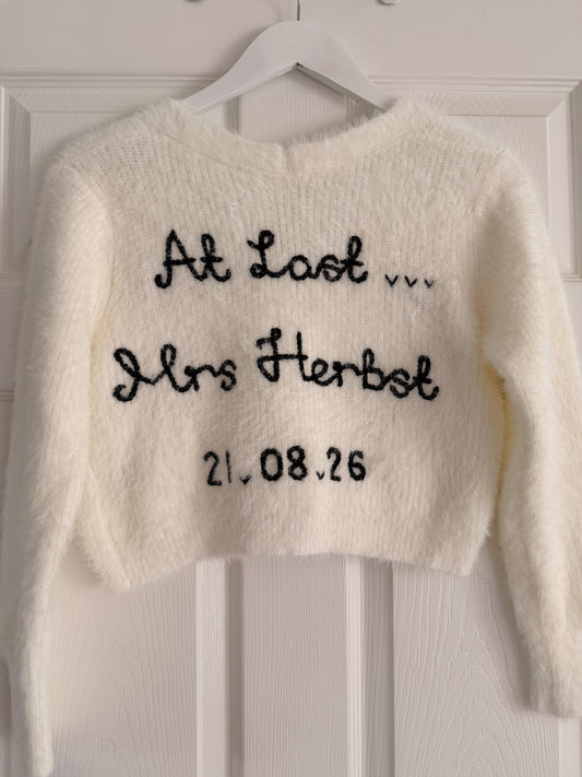 A fuzzy off-white cropped sweater hanging on a white hanger against a white paneled door. The sweater features black embroidered text reading 'At Last.