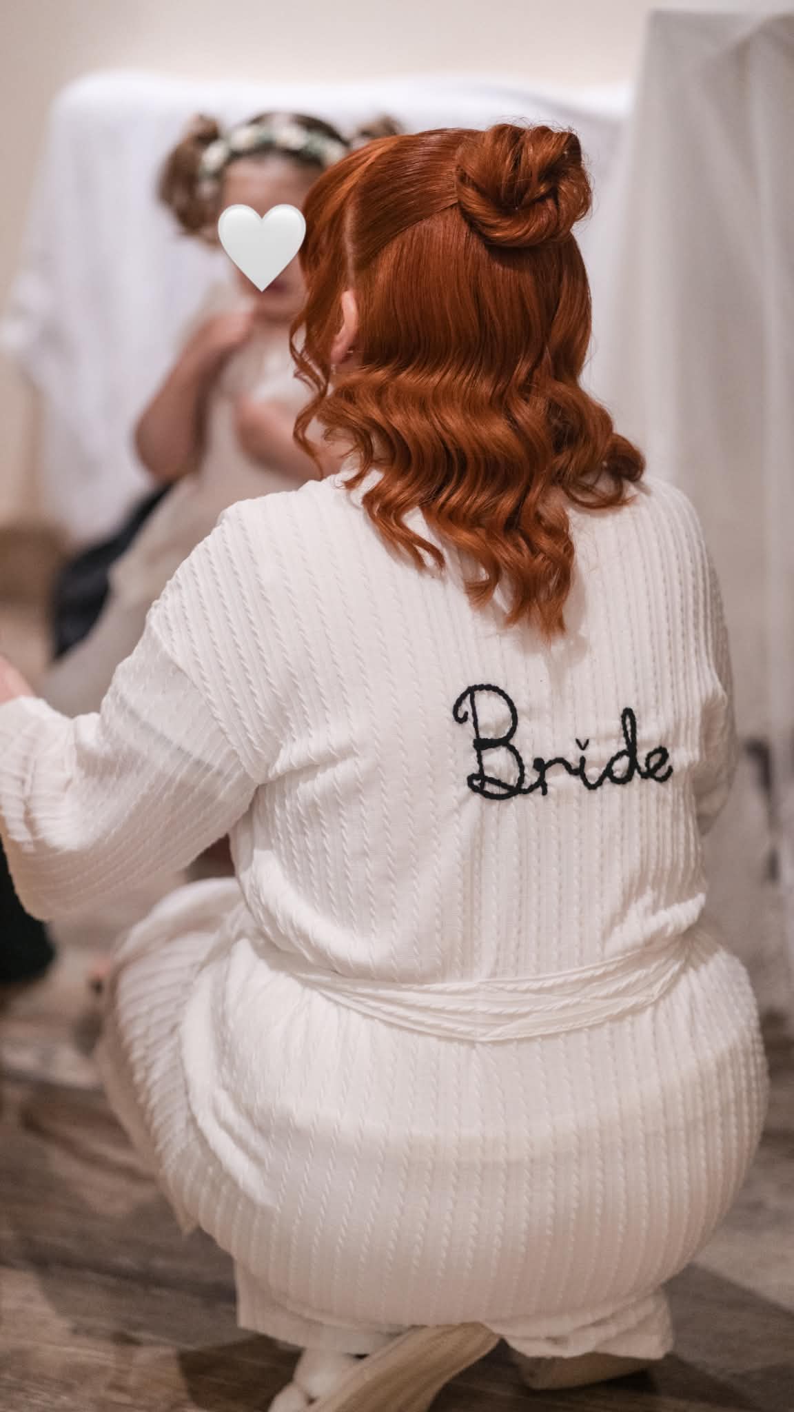 A woman with red hair seen from behind wearing a white ribbed robe with 'Bride' embroidered in black script on the back, kneeling in a bridal preparation setting.