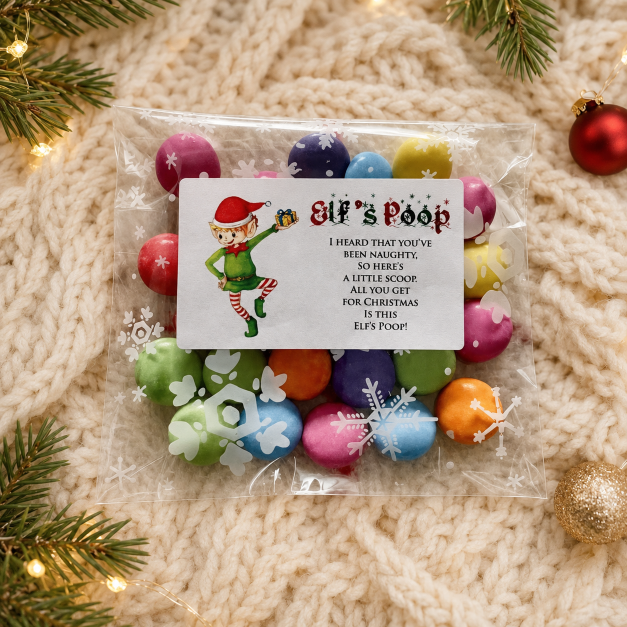 A clear plastic bag filled with colorful poop-shaped candies sits on a cream knit blanket, surrounded by pine branches and small holiday ornaments. The bag has a white printed label with an elf illustration and the text 'Elf's Poop' followed by a humorous Christmas message.