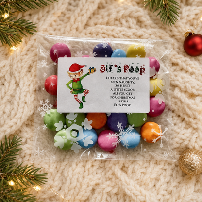 A clear plastic bag filled with colorful poop-shaped candies sits on a cream knit blanket, surrounded by pine branches and small holiday ornaments. The bag has a white printed label with an elf illustration and the text 'Elf's Poop' followed by a humorous Christmas message.