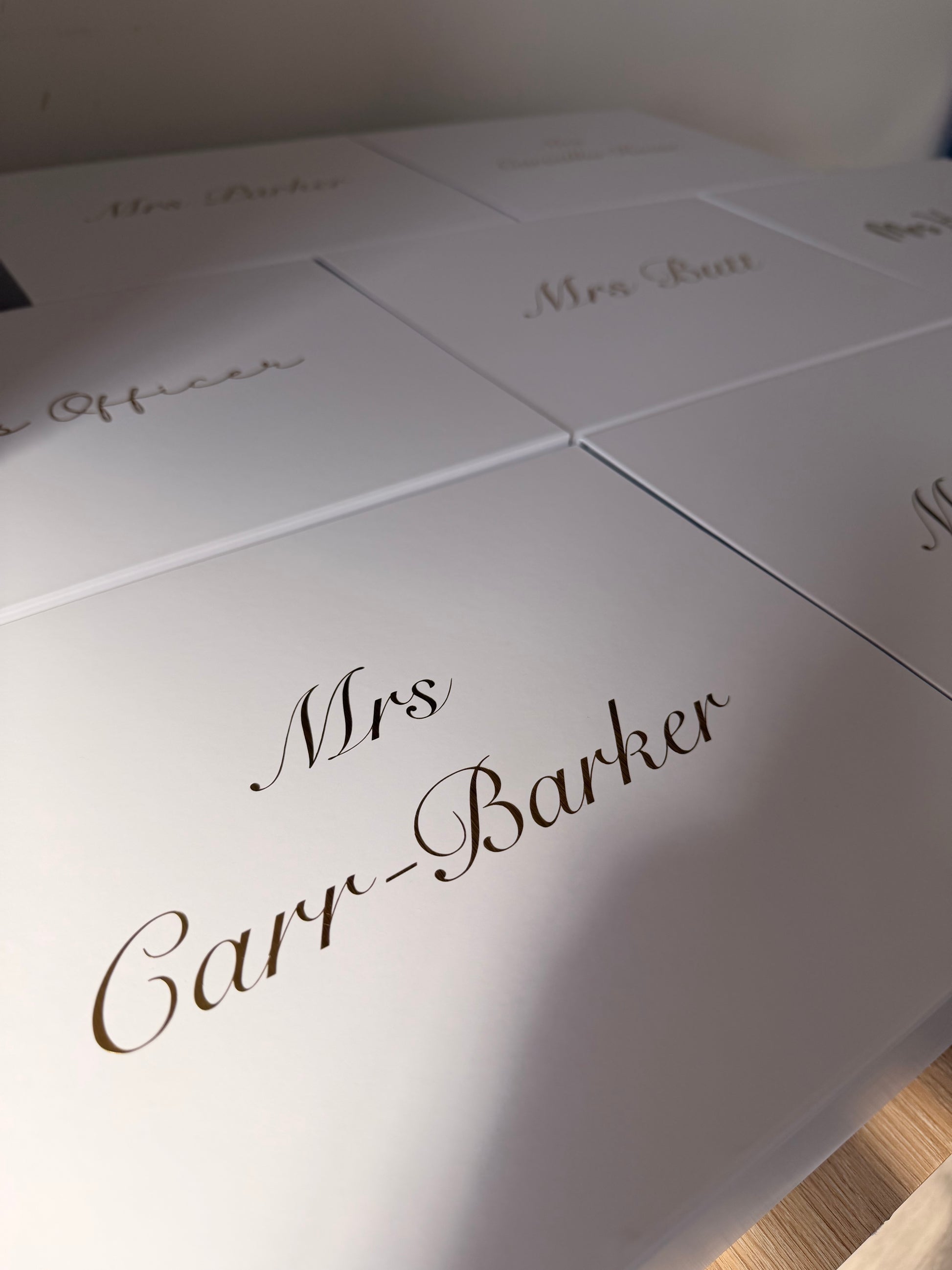 A close-up of a white personalized gift box or package lid with elegant cursive text reading 'Mrs. Cary-Barker' in dark brown or black script, with other similar boxes partially visible in the background.