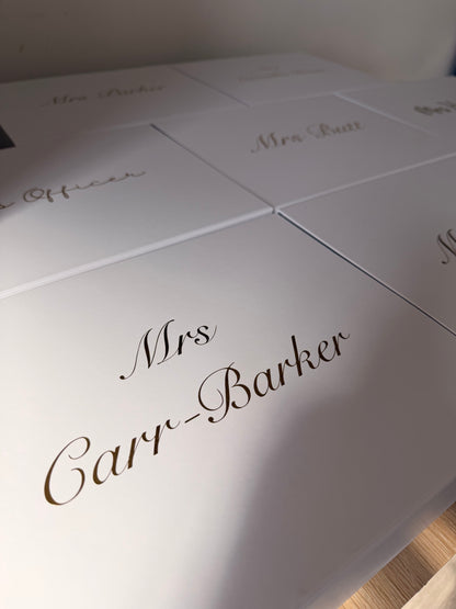 A close-up of a white personalized gift box or package lid with elegant cursive text reading 'Mrs. Cary-Barker' in dark brown or black script, with other similar boxes partially visible in the background.