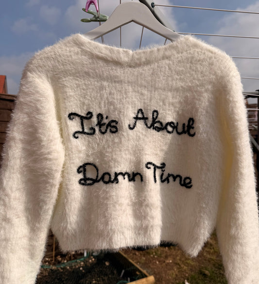 White fuzzy sweater with text 'It's About Damn Time' on a hanger outdoors.