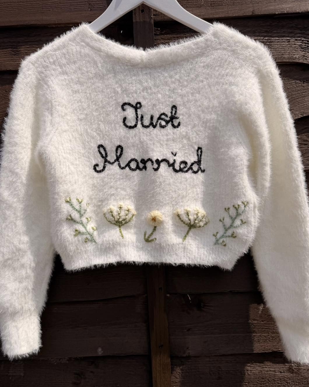 A white fluffy long-sleeve cropped cardigan hanging on a white hanger against a dark wooden plank background. The sweater features the words 'Just Married' embroidered in black script across the chest, with three small embroidered flowers and green stems below.