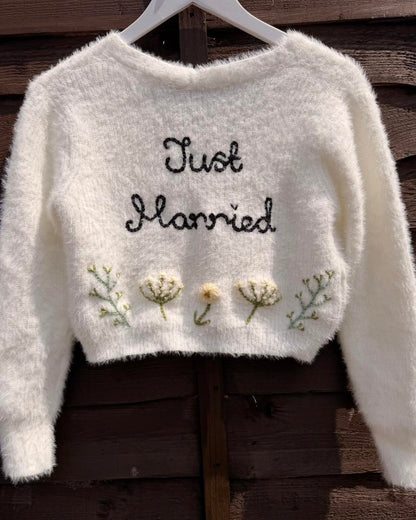 A white fluffy long-sleeve cropped cardigan hanging on a white hanger against a dark wooden plank background. The sweater features the words 'Just Married' embroidered in black script across the chest, with three small embroidered flowers and green stems below.