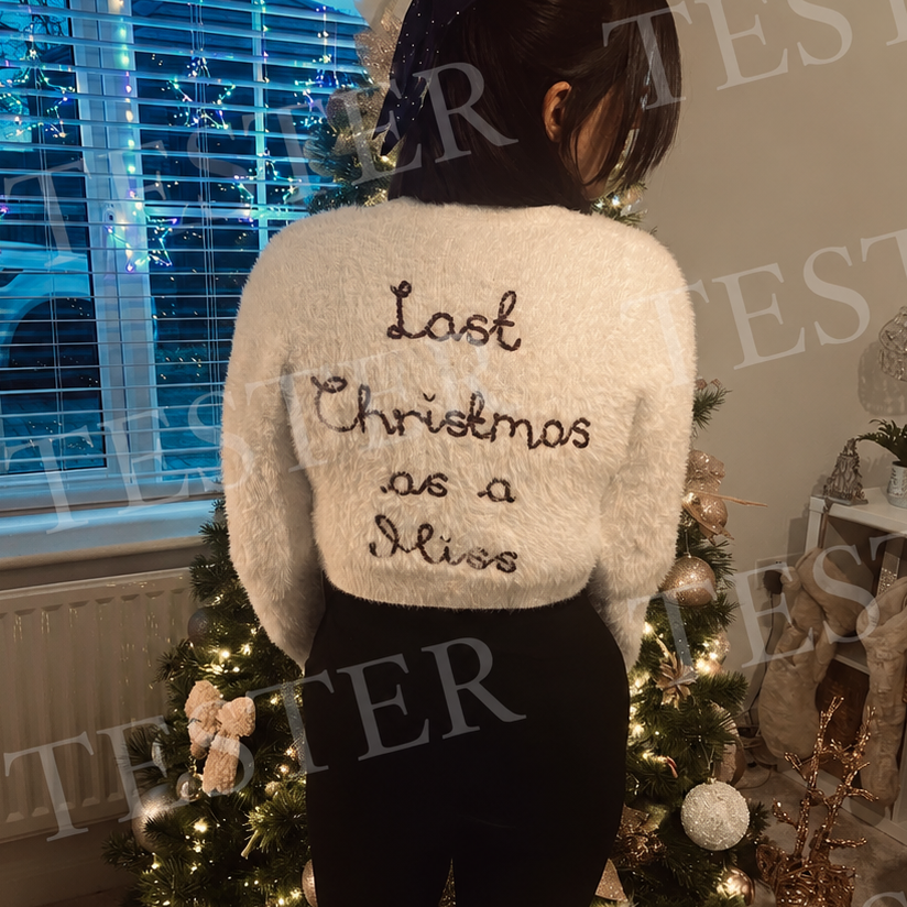 A woman viewed from behind wearing a fluffy white cropped sweater with the phrase 'Last Christmas as a Miss' embroidered in black across the back, standing in front of a decorated Christmas tree in a home interior.