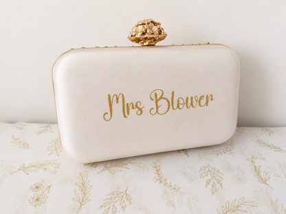 White clutch bag with 'Mrs Blower' text on a floral surface