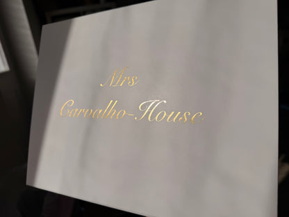 A close-up of a white box with elegant gold foil script reading 'Mrs Carvello-House' against a dark background.