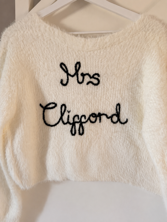 A white fluffy cropped cardigan with 'Mrs Clifford' embroidered in black script across the chest, hanging on a white hanger.