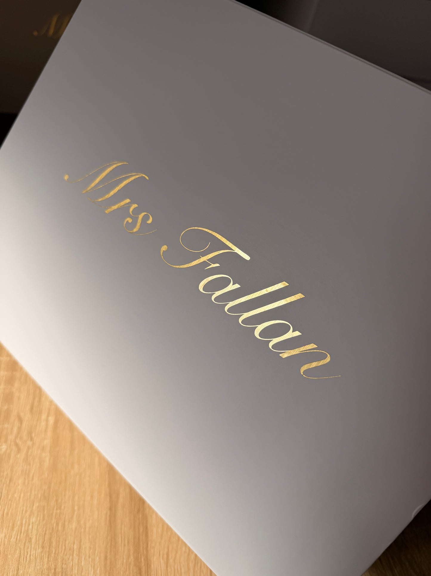 A close-up of a white rectangular box with elegant gold foil script reading 'Mrs. Fallon' on the lid, resting on a light wood surface.