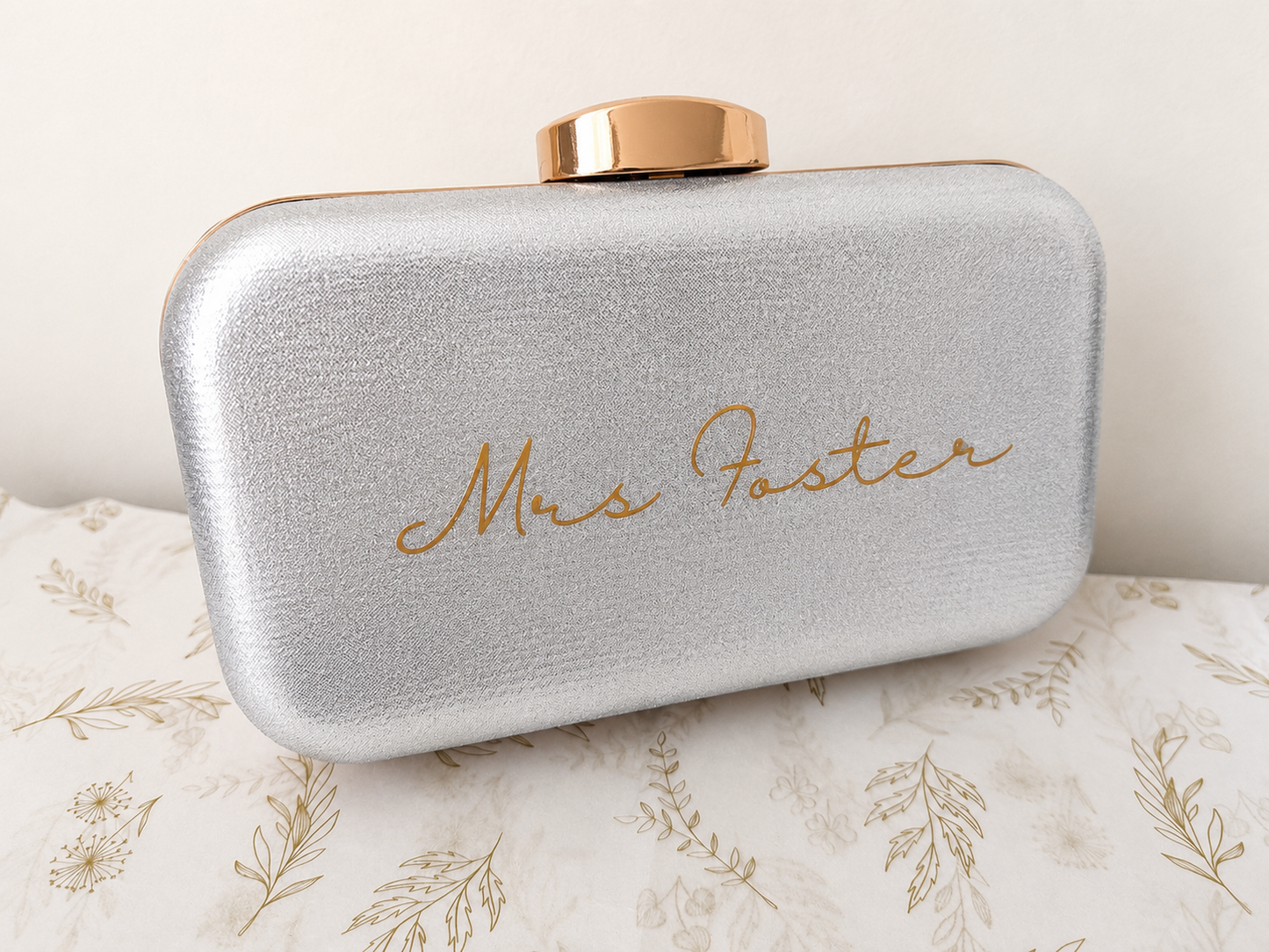 Silver clutch bag with 'Mrs. Foster' engraving on a floral-patterned surface