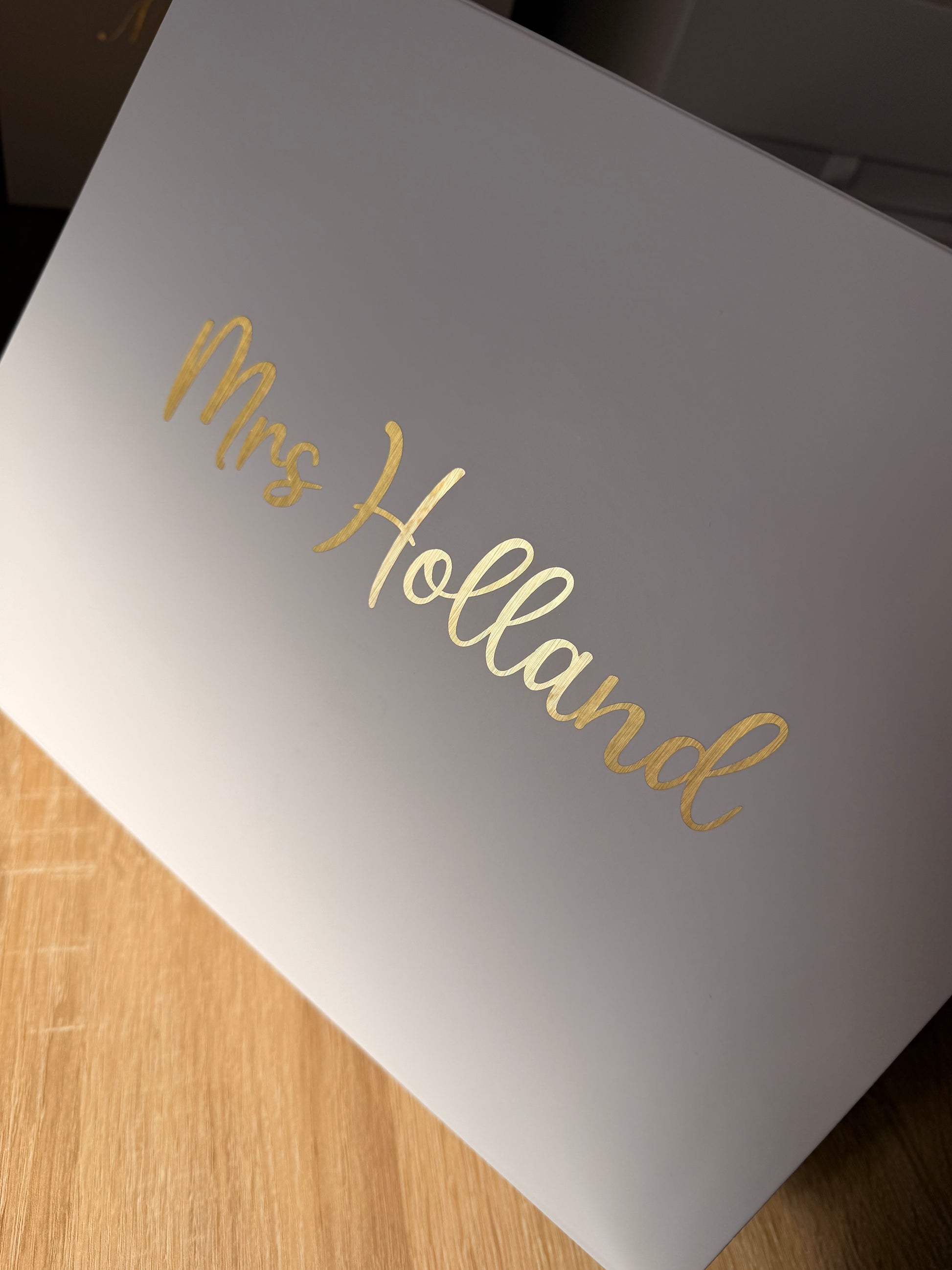 A white rectangular box with the name 'Mrs Holland' written in elegant gold cursive foil lettering, photographed at an angle on a light wood surface.
