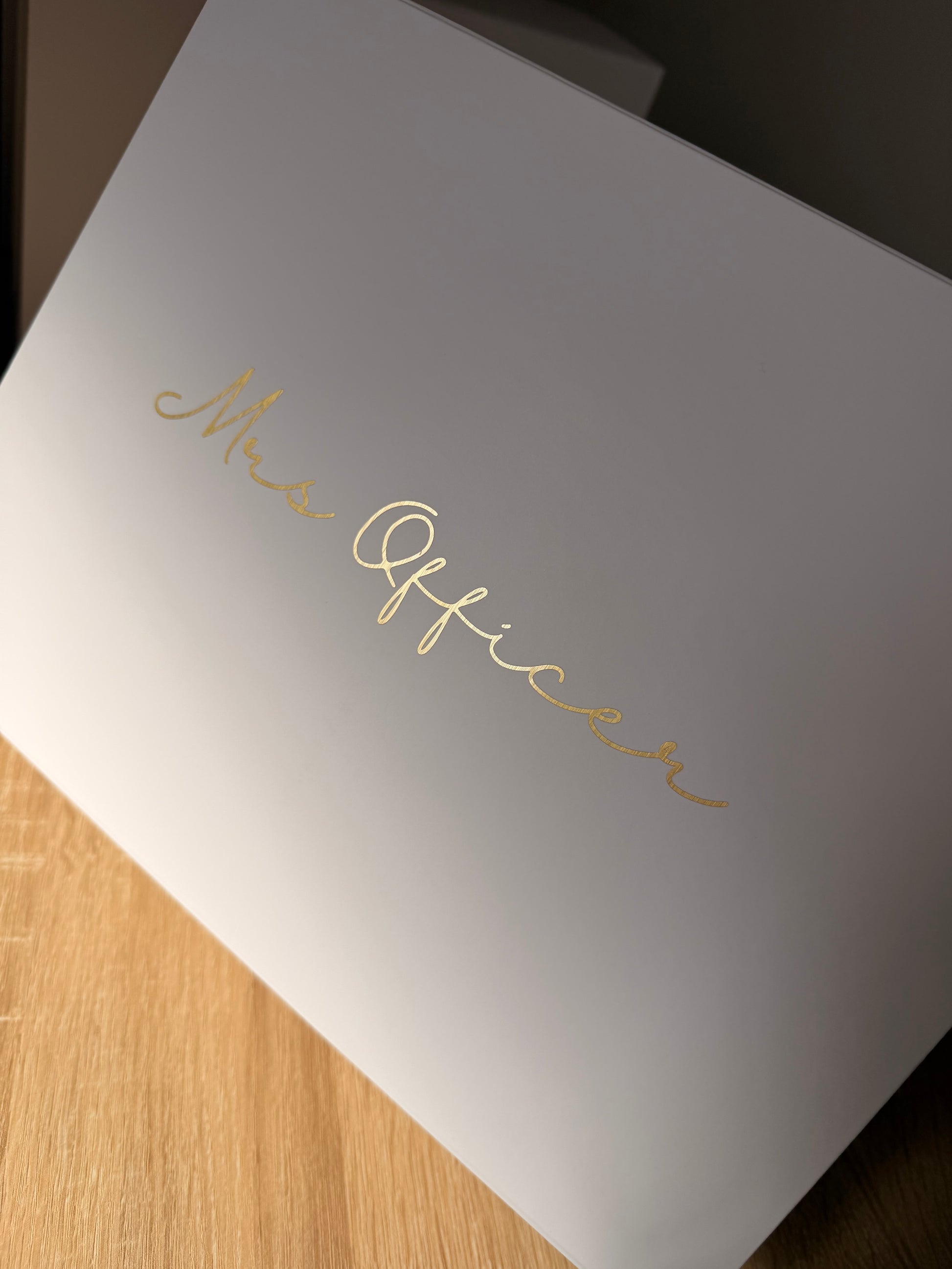 A white rectangular box with elegant gold foil script reading 'Mrs. Officer' on the lid, photographed at an angle on a light wood surface.
