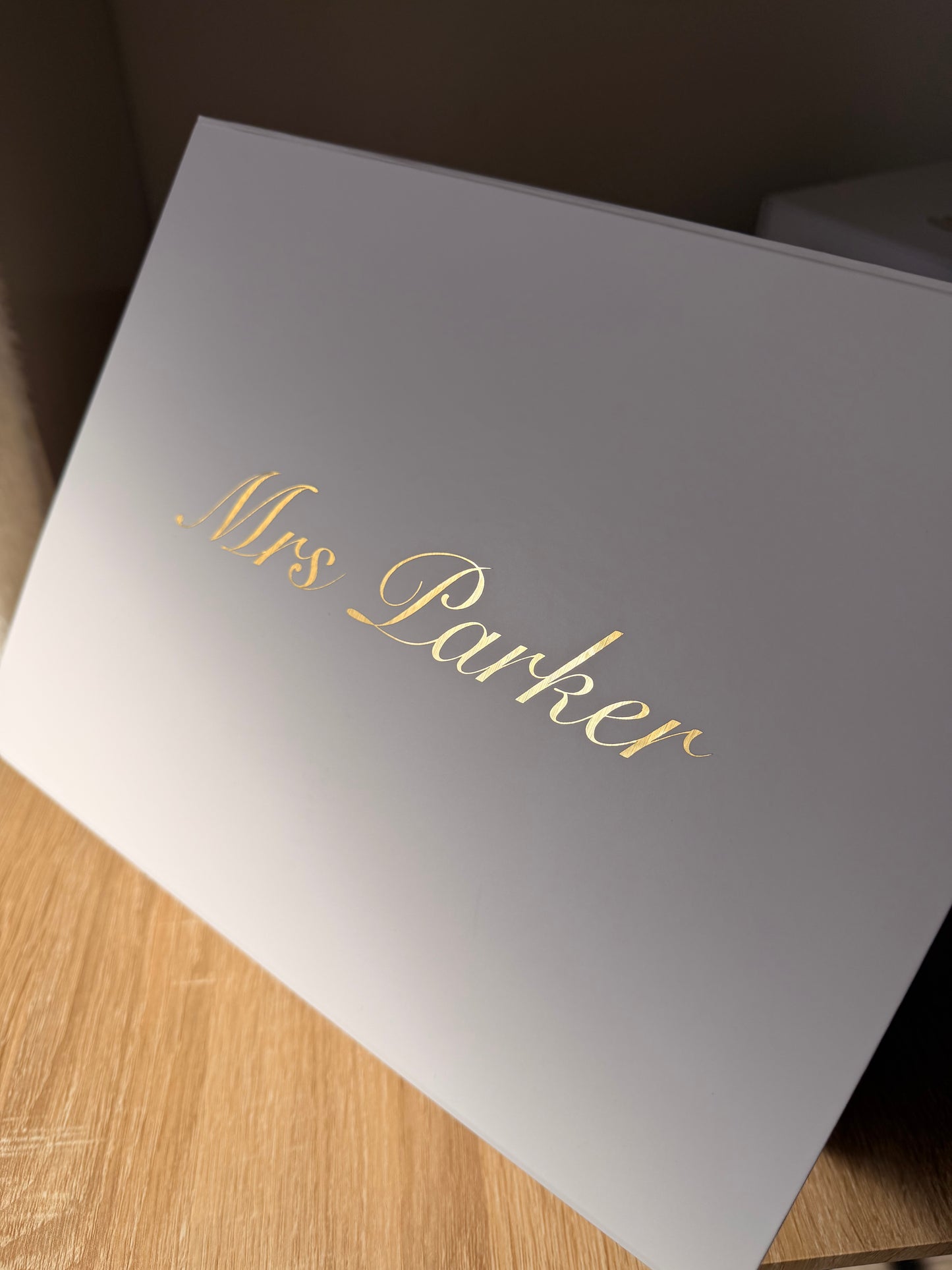 A white rectangular box with the name 'Mrs Parker' written in elegant gold cursive script, photographed at an angle on a light wood surface.