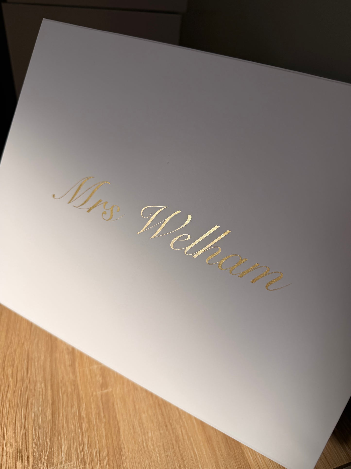 A white rectangular box with the name 'Mrs Welham' written in elegant gold cursive script, photographed at an angle on a light wood surface.