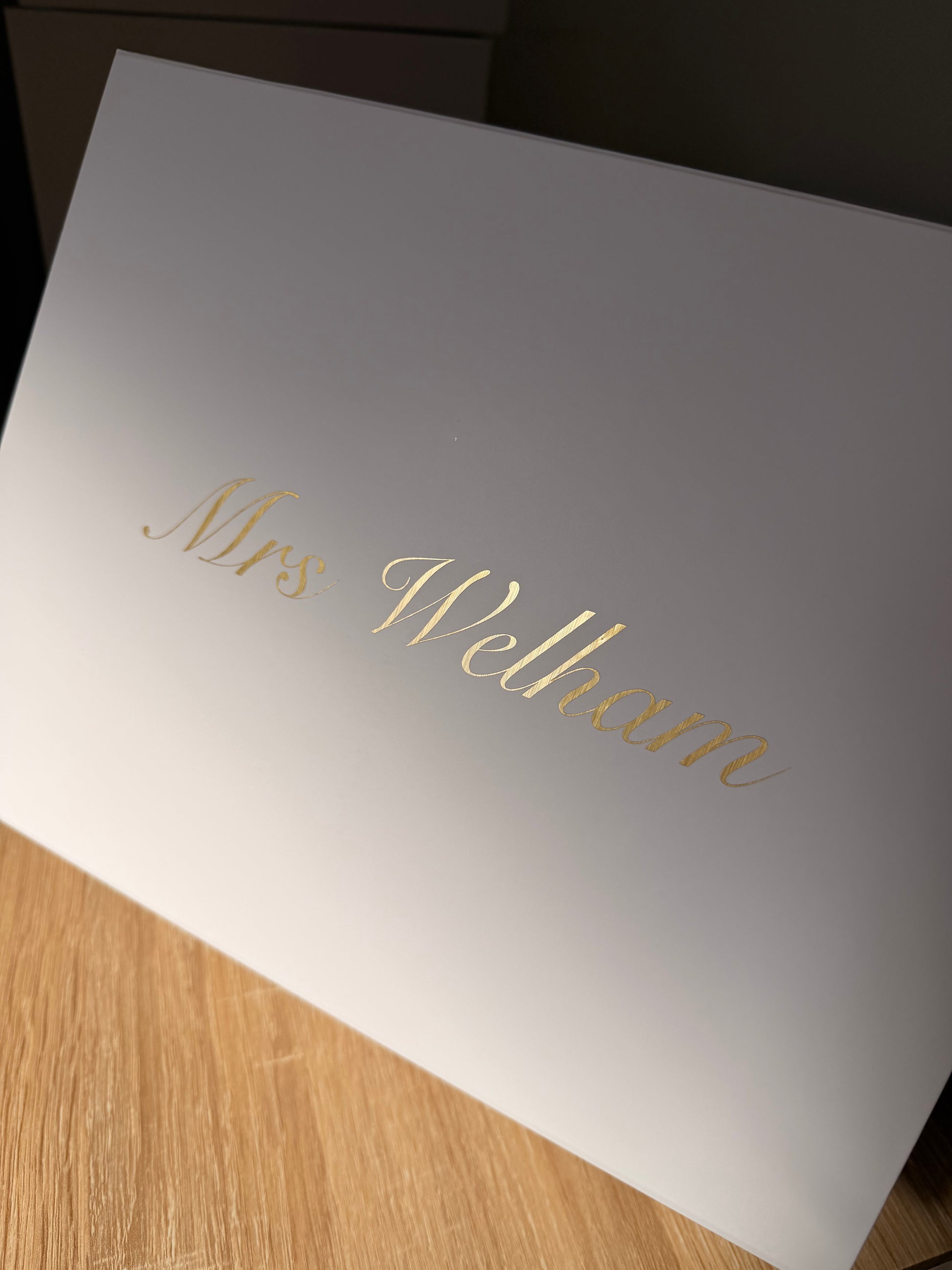 A white rectangular box with the name 'Mrs Welham' written in elegant gold cursive script, photographed at an angle on a light wood surface.
