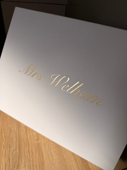 A white rectangular box with the name 'Mrs Welham' written in elegant gold cursive script, photographed at an angle on a light wood surface.
