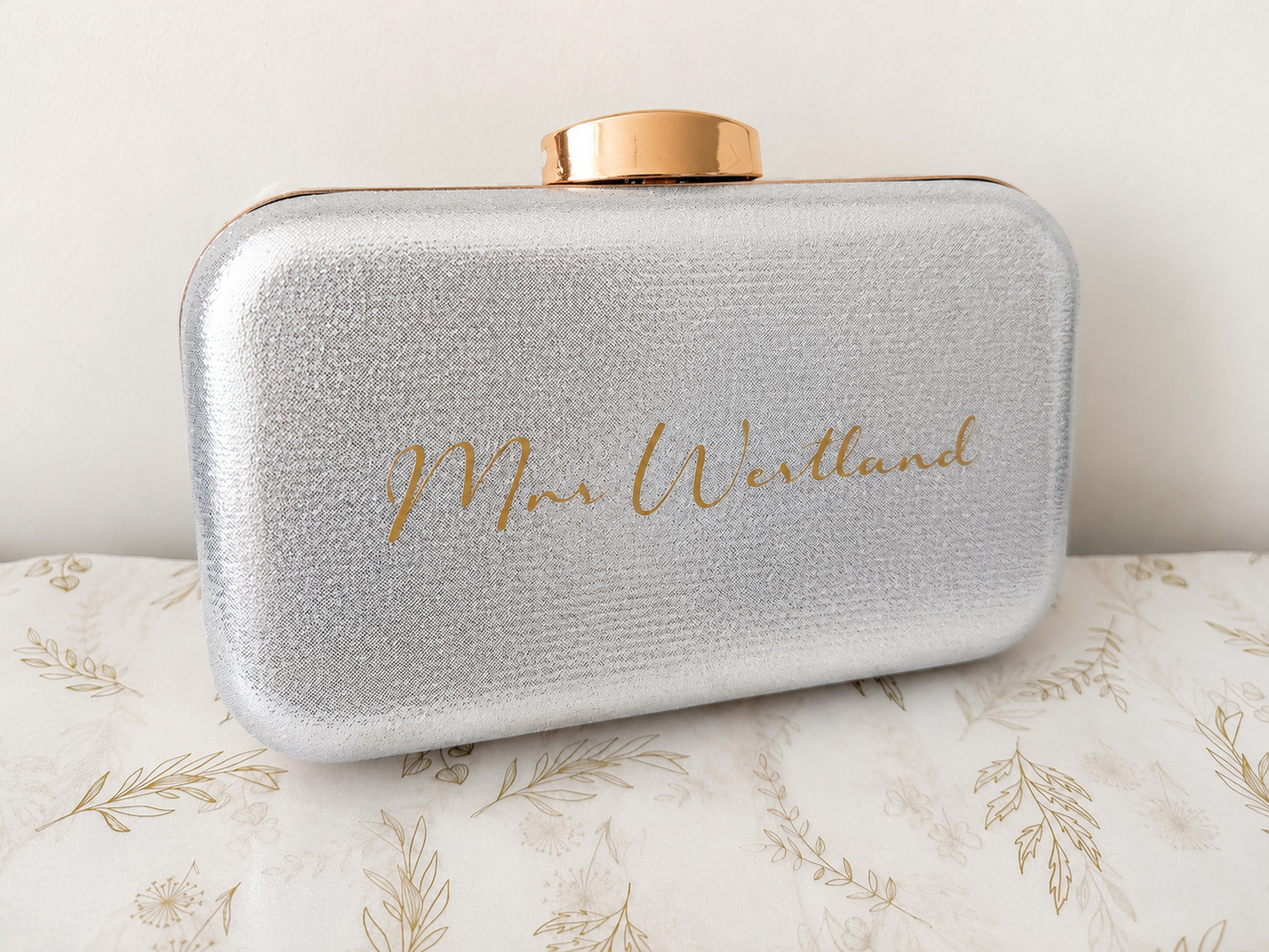 Silver clutch bag with gold lid and 'Mrs. Westlund' engraving on a floral-patterned surface.