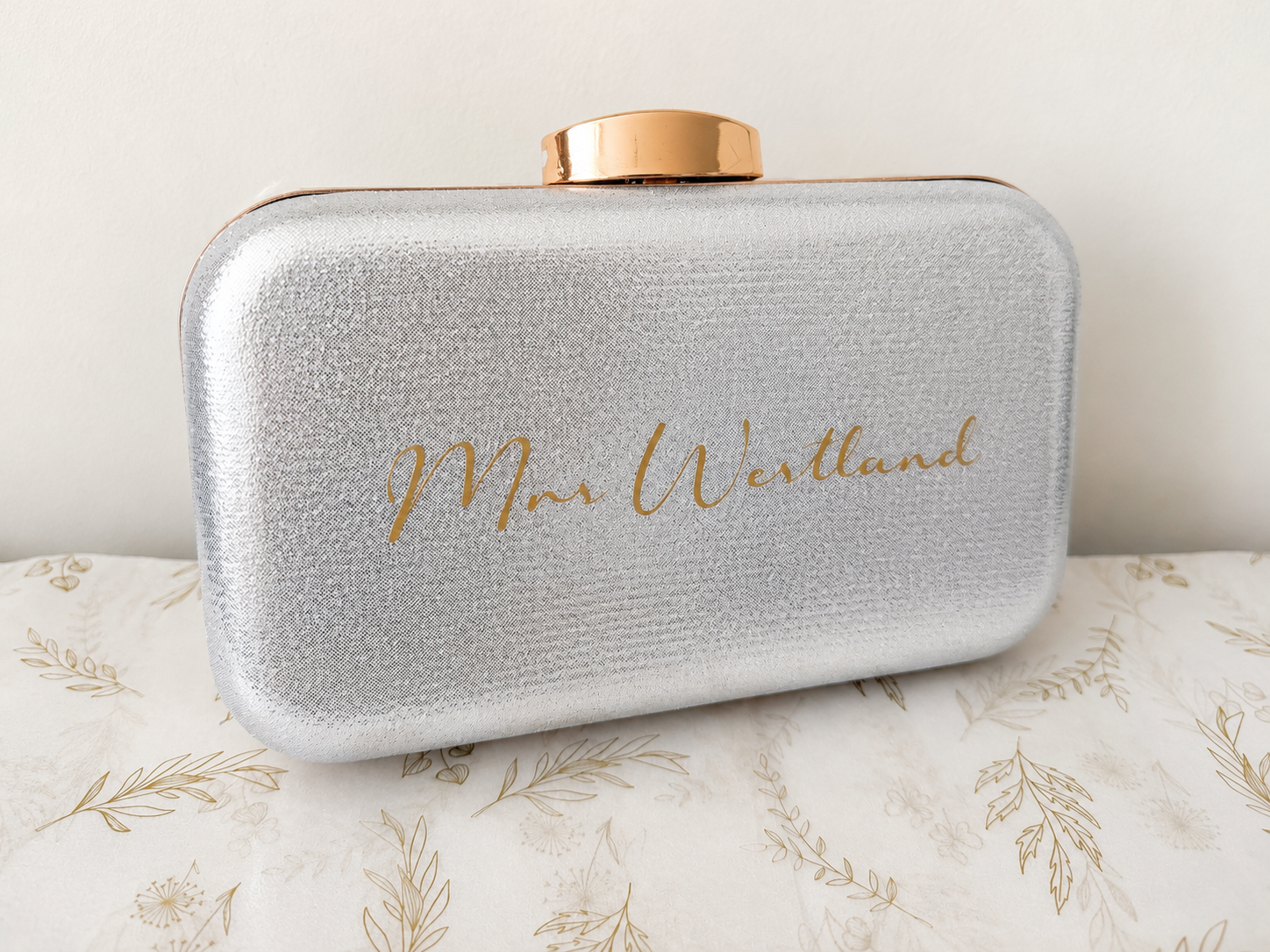 Silver clutch bag with gold lid and 'Mrs. Westlund' engraving on a floral-patterned surface.