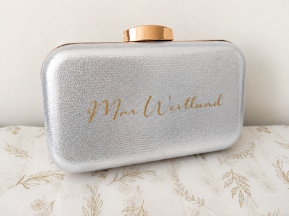 Silver clutch bag with gold lid and 'Mrs. Westlund' engraving on a floral-patterned surface.