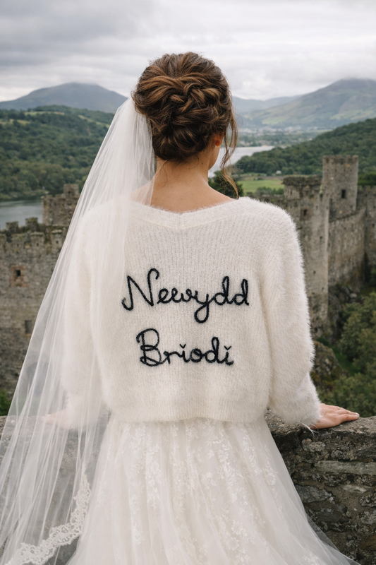 Woman in a wedding dress with 'Nevydd Briodi' text, standing in front of a castle.