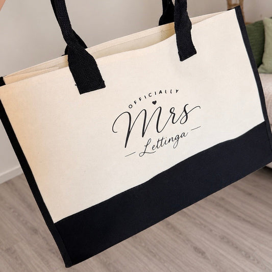 A cream canvas tote bag with black handles and a black bottom panel, printed with the text 'OFFICIALLY Mrs Lettinga' in black script. The bag is shown being held by a hand in a home setting with a light wood floor and a sofa in the background.