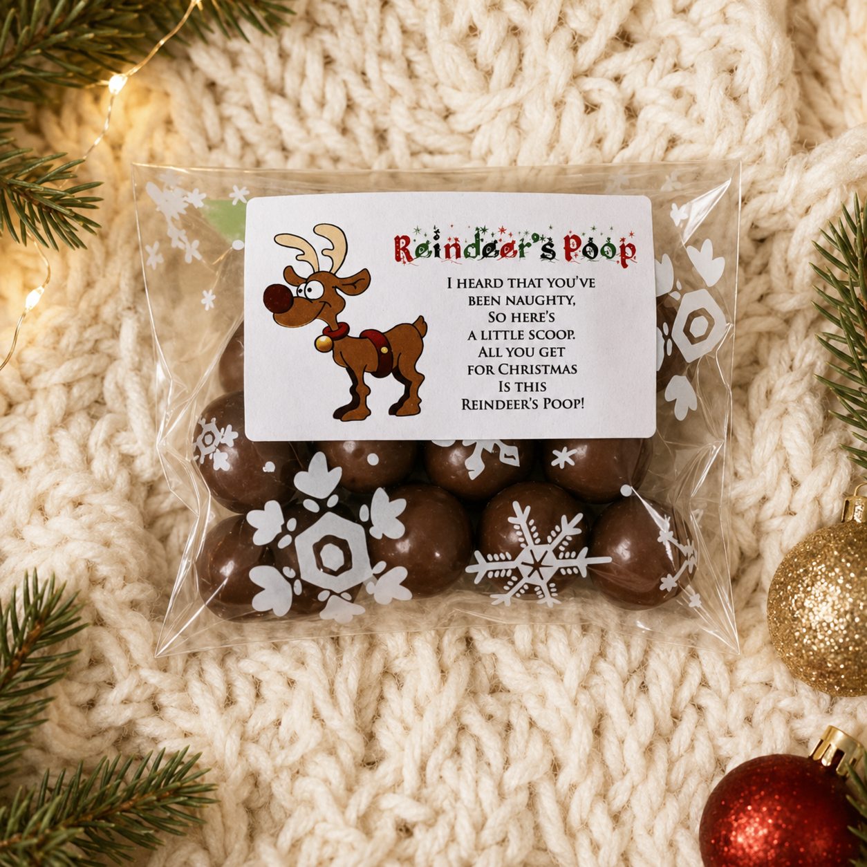 A clear plastic bag of chocolate treats filled with dark brown chocolates, sealed with a white printed label reading 'Reindeer's Poop' and featuring a cartoon reindeer and holiday text. The bag is photographed on a cream knit blanket with pine branches and Christmas ornaments around it.