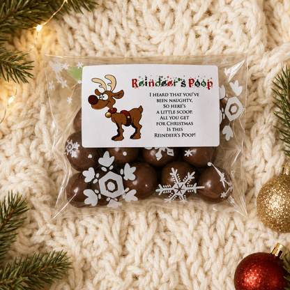 A clear plastic bag of chocolate treats filled with dark brown chocolates, sealed with a white printed label reading 'Reindeer's Poop' and featuring a cartoon reindeer and holiday text. The bag is photographed on a cream knit blanket with pine branches and Christmas ornaments around it.