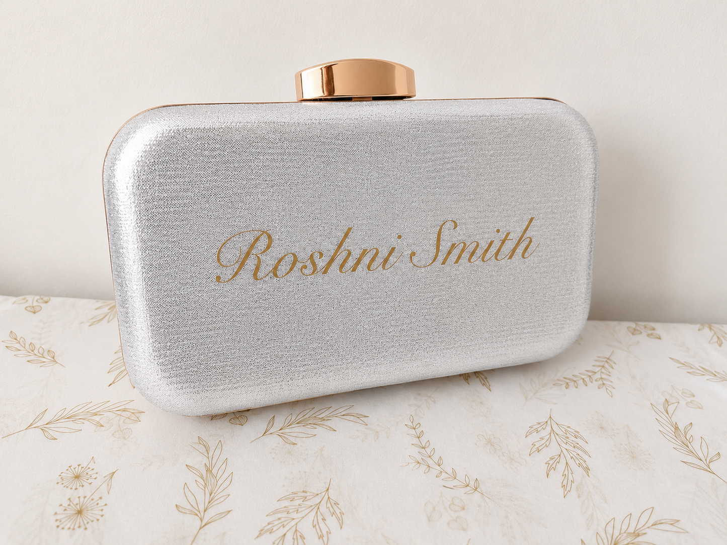 Silver clutch bag with gold text 'Roshni Smith' on a floral-patterned surface