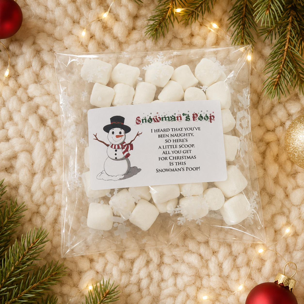 A clear plastic bag filled with mini marshmallows sits on a cream knit blanket with Christmas greenery and warm string lights. The bag has a white printed label that reads 'Snowman's Poop' with a snowman illustration and a humorous Christmas message.