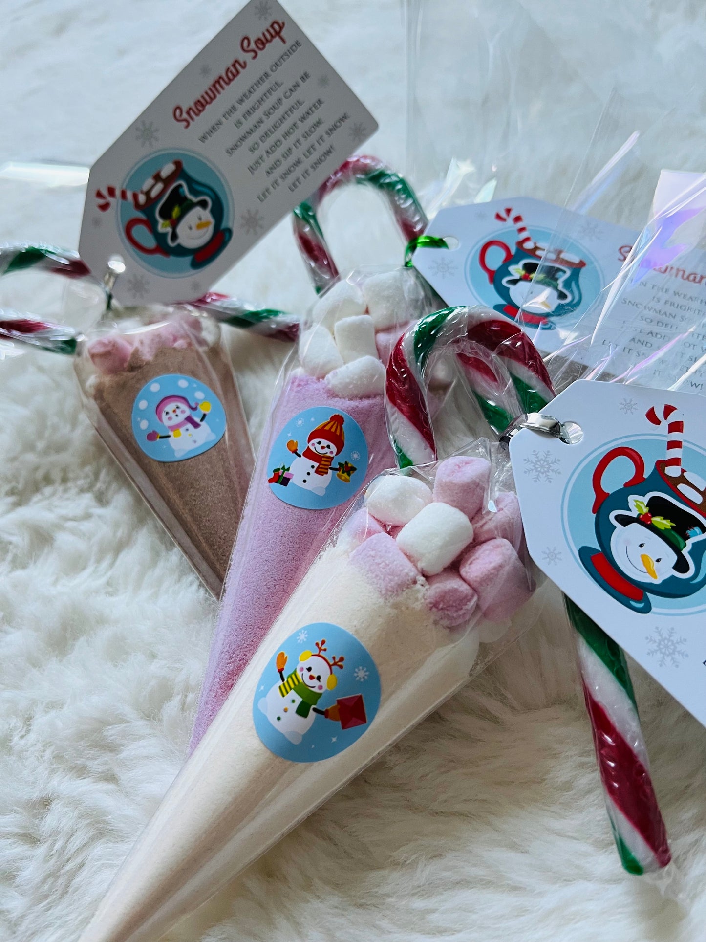Three small packaged holiday treats arranged on a white faux-fur surface. Each treat is wrapped in clear cellophane, tied with a candy cane, and has a round sticker with a penguin illustration.