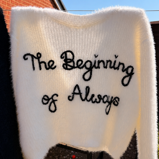 A white fuzzy knit sweater with the phrase 'The Beginning of Always' embroidered in black script across the chest, photographed outdoors against a brick wall and blue sky.