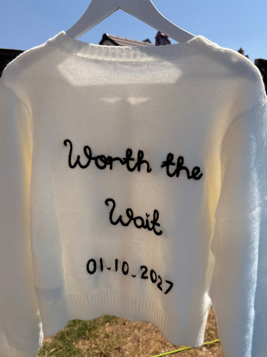 A white knit sweater hanging on a white hanger outdoors, with black handwritten text on the front reading 'Worth the Wait' and '01.10.
