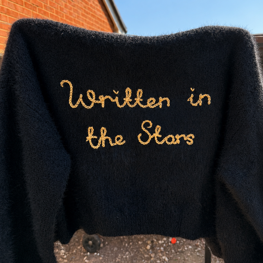 A black fuzzy knit sweater with 'Written in the Stars' embroidered in gold cursive text across the chest, photographed outdoors against a brick wall and blue sky.